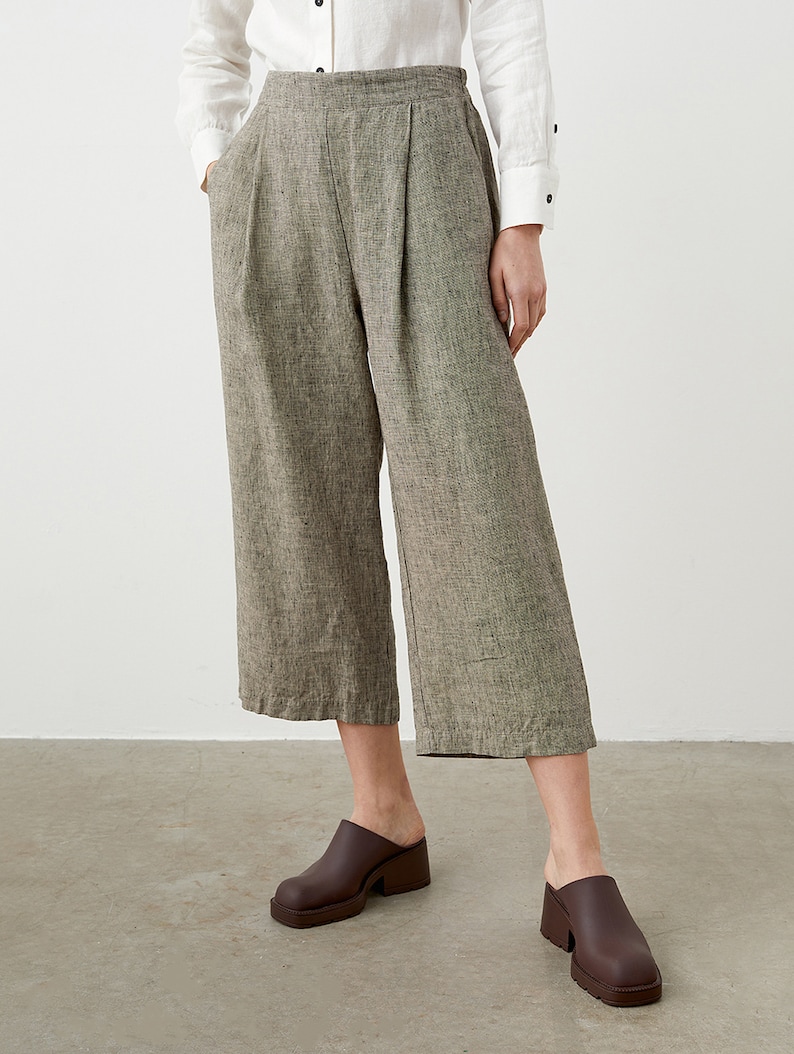 K&ouml;nnte beinhalten: Eine Hose mit weitem Bein und kurzem Schnitt aus strukturiertem, neutralem Stoff. Die Hose hat Falten an der Taille und Taschen. Das Model tr&auml;gt braune Clogs. Ein wei&szlig;es Hemd mit Kn&ouml;pfen ist teilweise sichtbar.