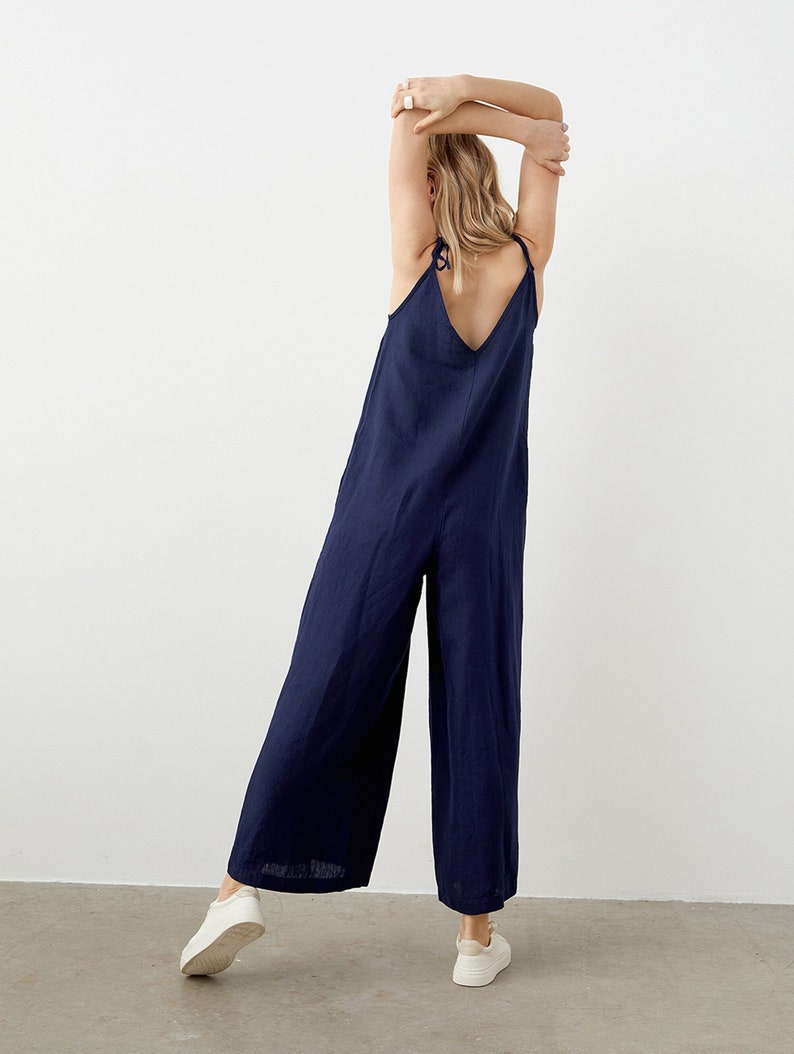 May include: A navy blue jumpsuit with wide legs and spaghetti straps. The jumpsuit has a V-neckline and tie details at the shoulders. The model is wearing white sneakers. The fabric appears to be linen or a similar lightweight material.