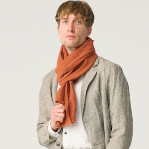 May include: A rust-colored scarf draped around the neck of a person wearing a light gray blazer and matching pants. The scarf is made of a soft, flowing fabric. The blazer has a classic two-button design and a black belt.