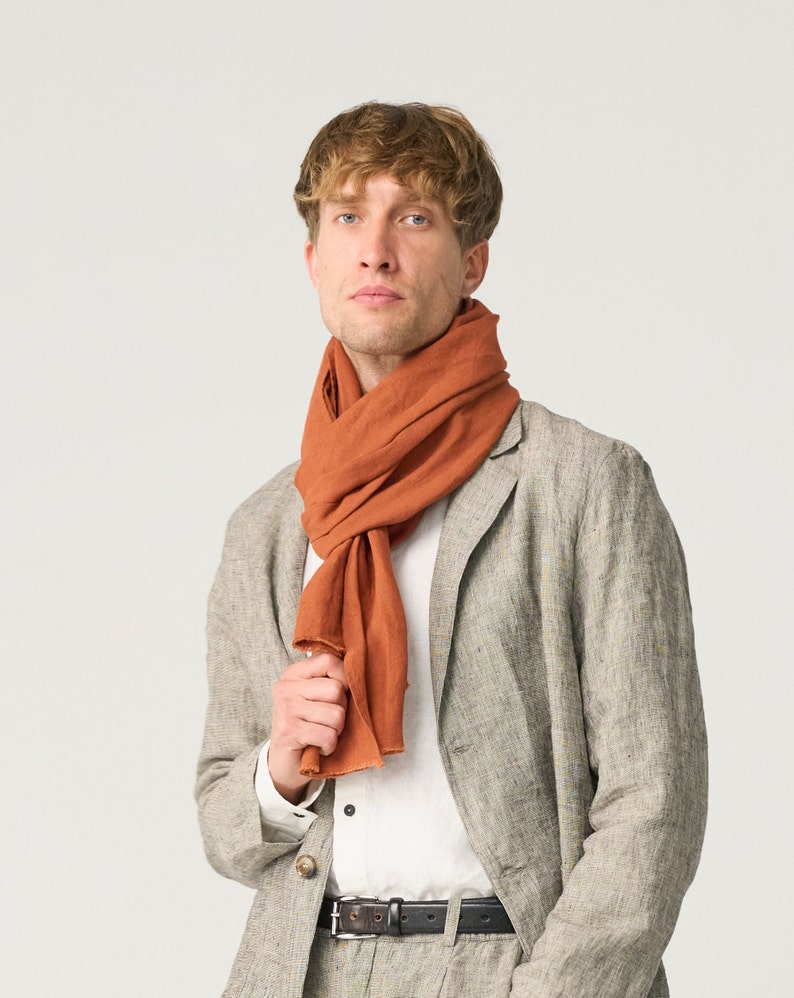 May include: A rust-colored scarf draped around the neck of a person wearing a light gray blazer and matching pants. The scarf is made of a soft, flowing fabric. The blazer has a classic two-button design and a black belt.