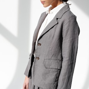 May include: A gray blazer with two buttons and a pocket, paired with matching pants. The blazer is worn over a white collared shirt. The outfit is made of a textured fabric, suggesting linen or a similar material. The overall style is classic and tailored.