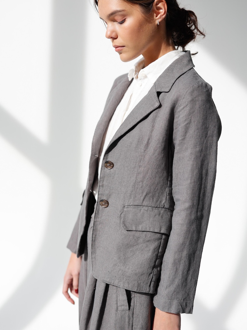 May include: A gray blazer with two buttons and a pocket, paired with matching pants. The blazer is worn over a white collared shirt. The outfit is made of a textured fabric, suggesting linen or a similar material. The overall style is classic and tailored.