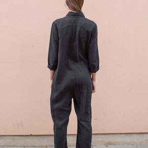 May include: A black linen jumpsuit with long sleeves and a collared neckline. The jumpsuit has a relaxed fit and is shown on a model standing in front of a pink wall.