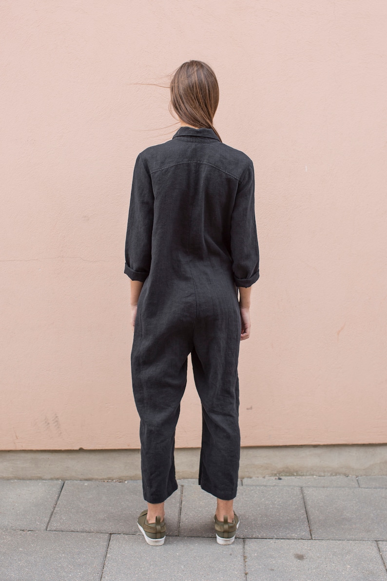 May include: A black linen jumpsuit with long sleeves and a collared neckline. The jumpsuit has a relaxed fit and is shown on a model standing in front of a pink wall.