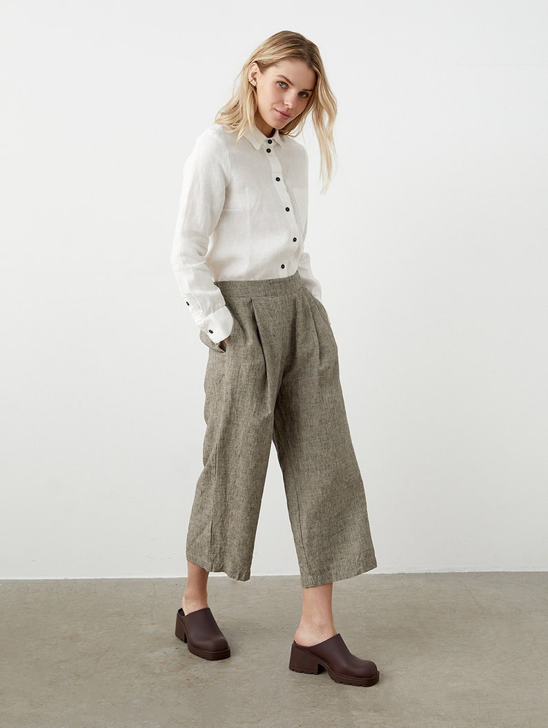 K&ouml;nnte beinhalten: Eine Frau tr&auml;gt ein wei&szlig;es Hemd mit schwarzen Kn&ouml;pfen und Manschetten, kombiniert mit einer weit geschnittenen, cropped Hose in Grau-Braun. Sie tr&auml;gt braune Lederclogs. Das Outfit ist vor einem schlichten wei&szlig;en Hintergrund abgebildet.