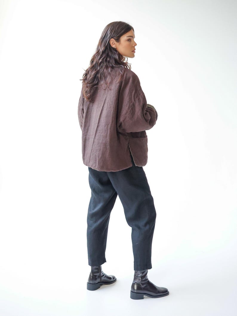 May include: A person wearing a brown linen jacket, black trousers, and black ankle boots. The jacket has a relaxed fit and a simple design. The trousers are a dark colour and have a slightly loose fit. The boots are leather.