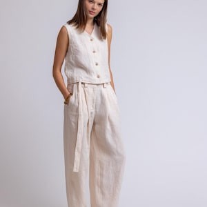 May include: A two-piece linen outfit in a neutral beige color. The sleeveless top has a V-neck and wooden buttons. The high-waisted pants have a matching belt. The outfit is made of a lightweight fabric.