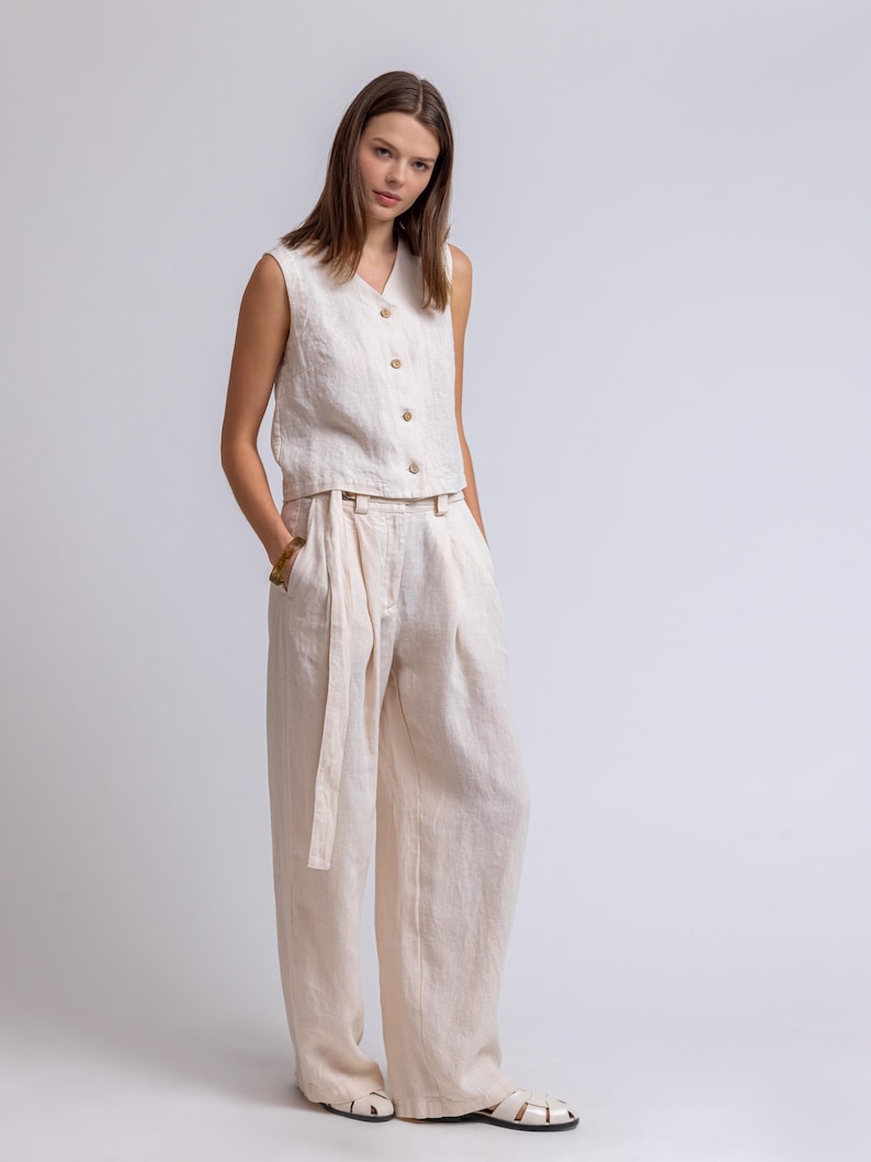 May include: A two-piece linen outfit in a neutral beige color. The sleeveless top has a V-neck and wooden buttons. The high-waisted pants have a matching belt. The outfit is made of a lightweight fabric.
