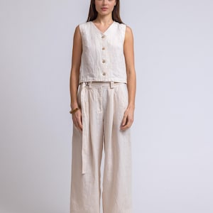 May include: A cream-colored linen two-piece outfit. The sleeveless top has a V-neck and wooden buttons. The high-waisted, wide-leg pants have a matching fabric belt. The outfit is made of a lightweight, breathable fabric, perfect for warm weather.
