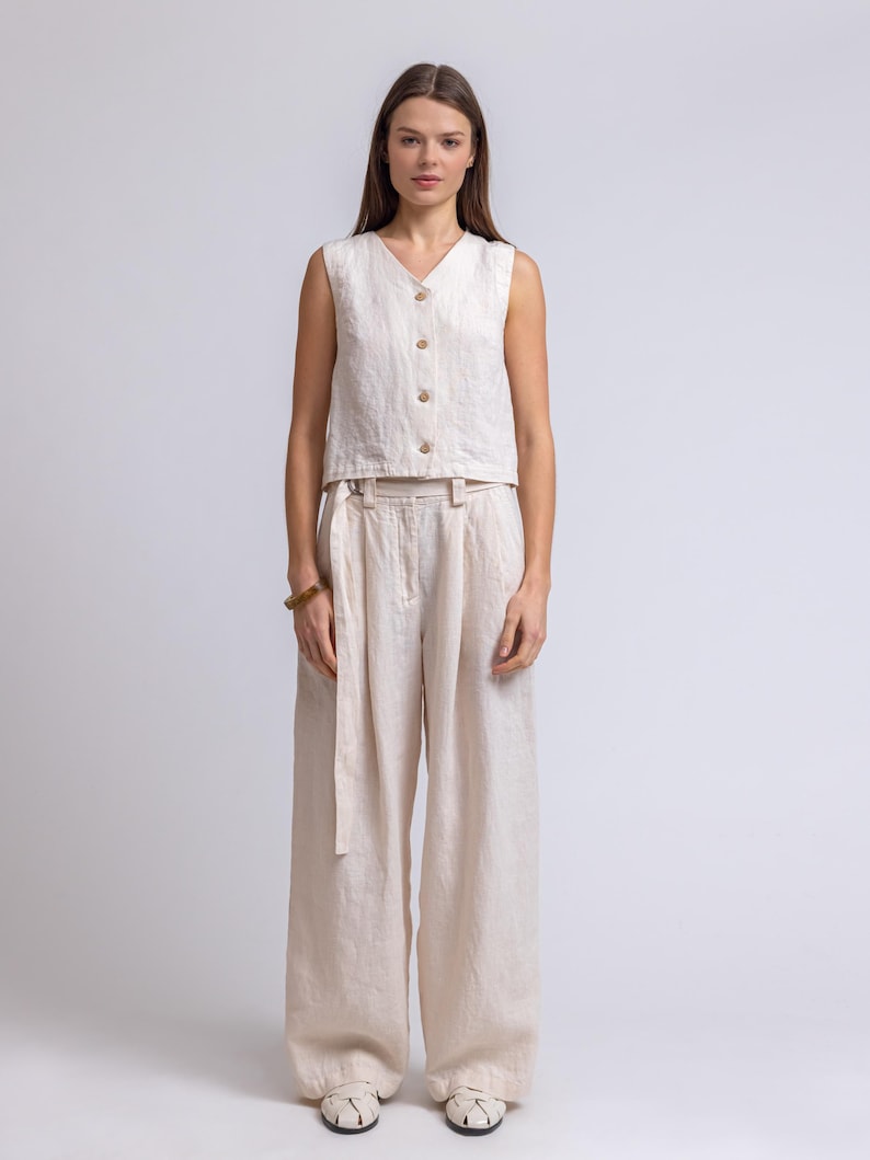 May include: A cream-colored linen two-piece outfit. The sleeveless top has a V-neck and wooden buttons. The high-waisted, wide-leg pants have a matching fabric belt. The outfit is made of a lightweight, breathable fabric, perfect for warm weather.