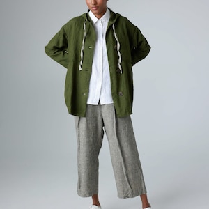 K&ouml;nnte beinhalten: Eine Person tr&auml;gt eine gr&uuml;ne Kapuzenjacke, ein wei&szlig;es Hemd und eine graue Leinenhose. Die Jacke hat eine Kapuze mit Kordelzug und Holzkn&ouml;pfe. Wei&szlig;e Turnschuhe vervollst&auml;ndigen das Outfit. Die Kleidung ist aus Leinen.