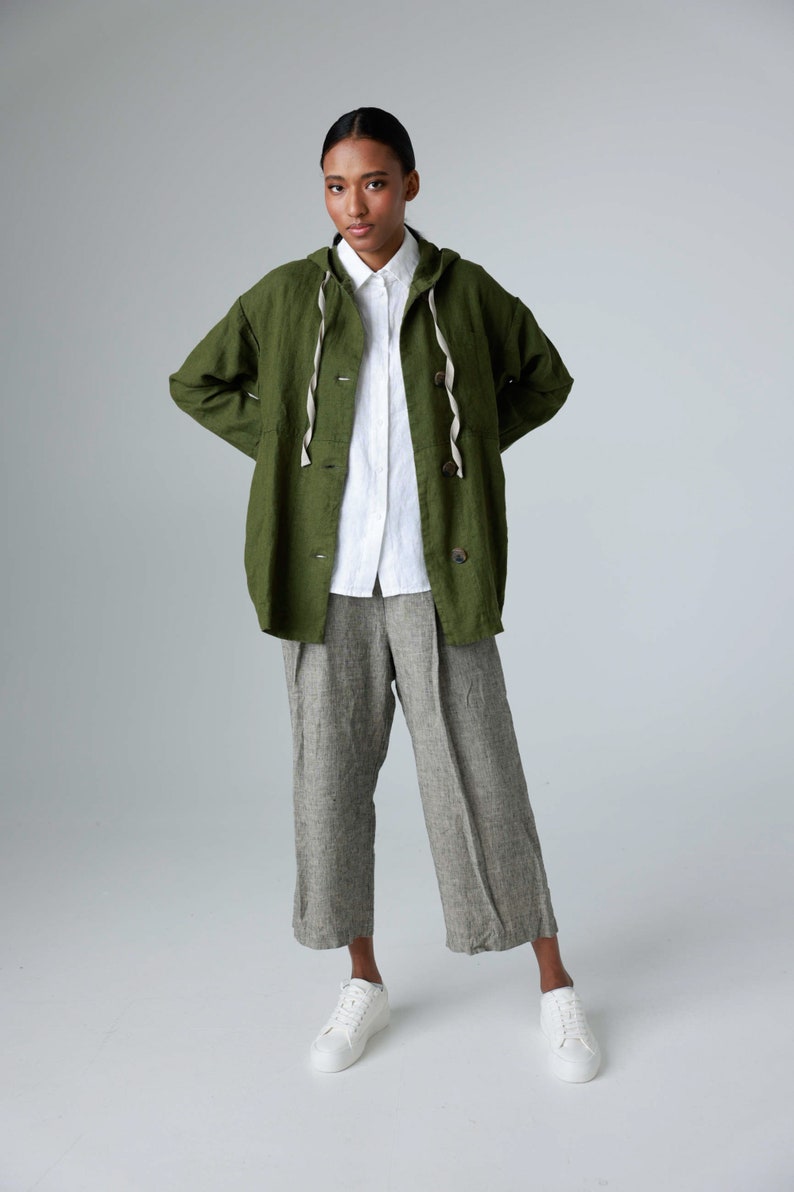 K&ouml;nnte beinhalten: Eine Person tr&auml;gt eine gr&uuml;ne Kapuzenjacke, ein wei&szlig;es Hemd und eine graue Leinenhose. Die Jacke hat eine Kapuze mit Kordelzug und Holzkn&ouml;pfe. Wei&szlig;e Turnschuhe vervollst&auml;ndigen das Outfit. Die Kleidung ist aus Leinen.