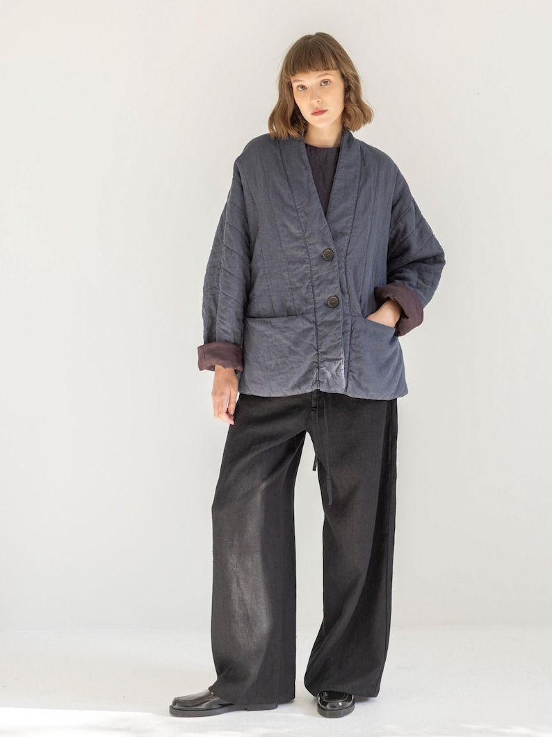 May include: A navy blue quilted jacket with a V-neck, two large buttons, and two front pockets. The jacket has a brown lining and is paired with black wide-leg trousers. The outfit is simple and elegant.