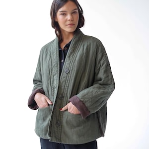 May include: An olive green quilted jacket with a relaxed fit, button-up front, and two front pockets. The jacket has brown cuffs and a collar. The model is wearing a black shirt and black trousers.