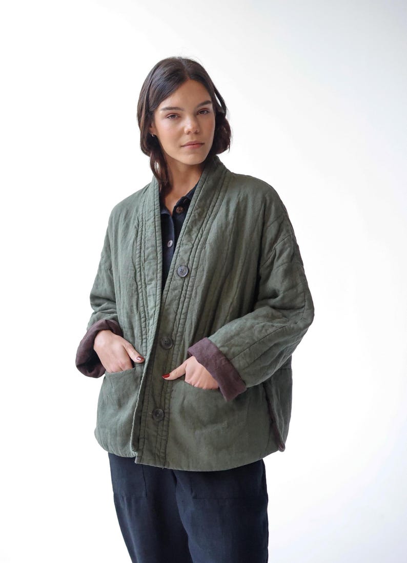 May include: An olive green quilted jacket with a relaxed fit, button-up front, and two front pockets. The jacket has brown cuffs and a collar. The model is wearing a black shirt and black trousers.
