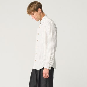 Op de afbeelding: Een wit overhemd met verticale zwarte strepen en houten knopen. Het shirt met lange mouwen wordt gecombineerd met een zwarte broek. Het model draagt het overhemd en de broek.