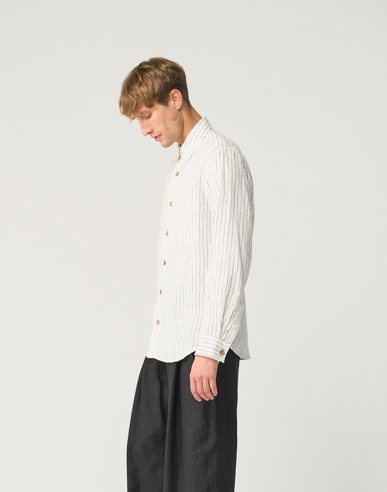 Op de afbeelding: Een wit overhemd met verticale zwarte strepen en houten knopen. Het shirt met lange mouwen wordt gecombineerd met een zwarte broek. Het model draagt het overhemd en de broek.