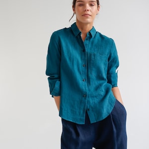 May include: Teal button-down shirt with a chest pocket and long sleeves, paired with navy blue pants. The shirt is made of a soft, flowing fabric. The model is wearing the shirt and pants.