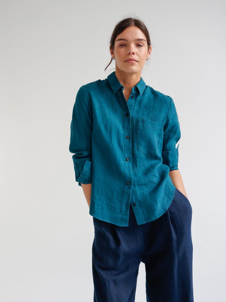 May include: Teal button-down shirt with a chest pocket and long sleeves, paired with navy blue pants. The shirt is made of a soft, flowing fabric. The model is wearing the shirt and pants.