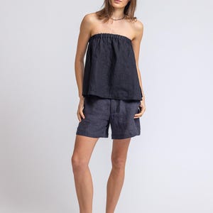 May include: A woman wearing a black strapless top and dark blue shorts. The top is gathered at the neckline. She is also wearing dark brown leather sandals. The outfit is made of a lightweight fabric.