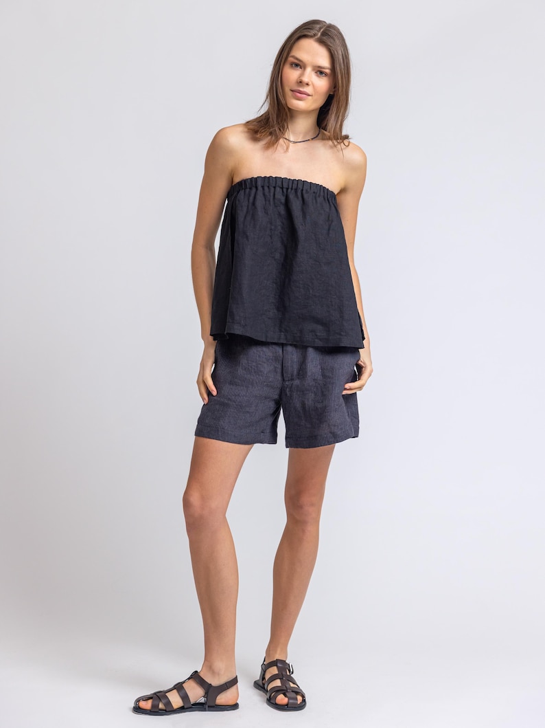 May include: A woman wearing a black strapless top and dark blue shorts. The top is gathered at the neckline. She is also wearing dark brown leather sandals. The outfit is made of a lightweight fabric.
