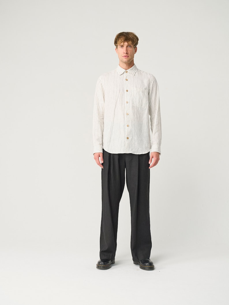 Op de afbeelding: Een wit overhemd met grijze verticale strepen en houten knopen, gecombineerd met een donkergrijze plooibroek en zwarte schoenen. De outfit wordt gepresenteerd tegen een witte achtergrond, wat een minimalistische esthetiek benadrukt.