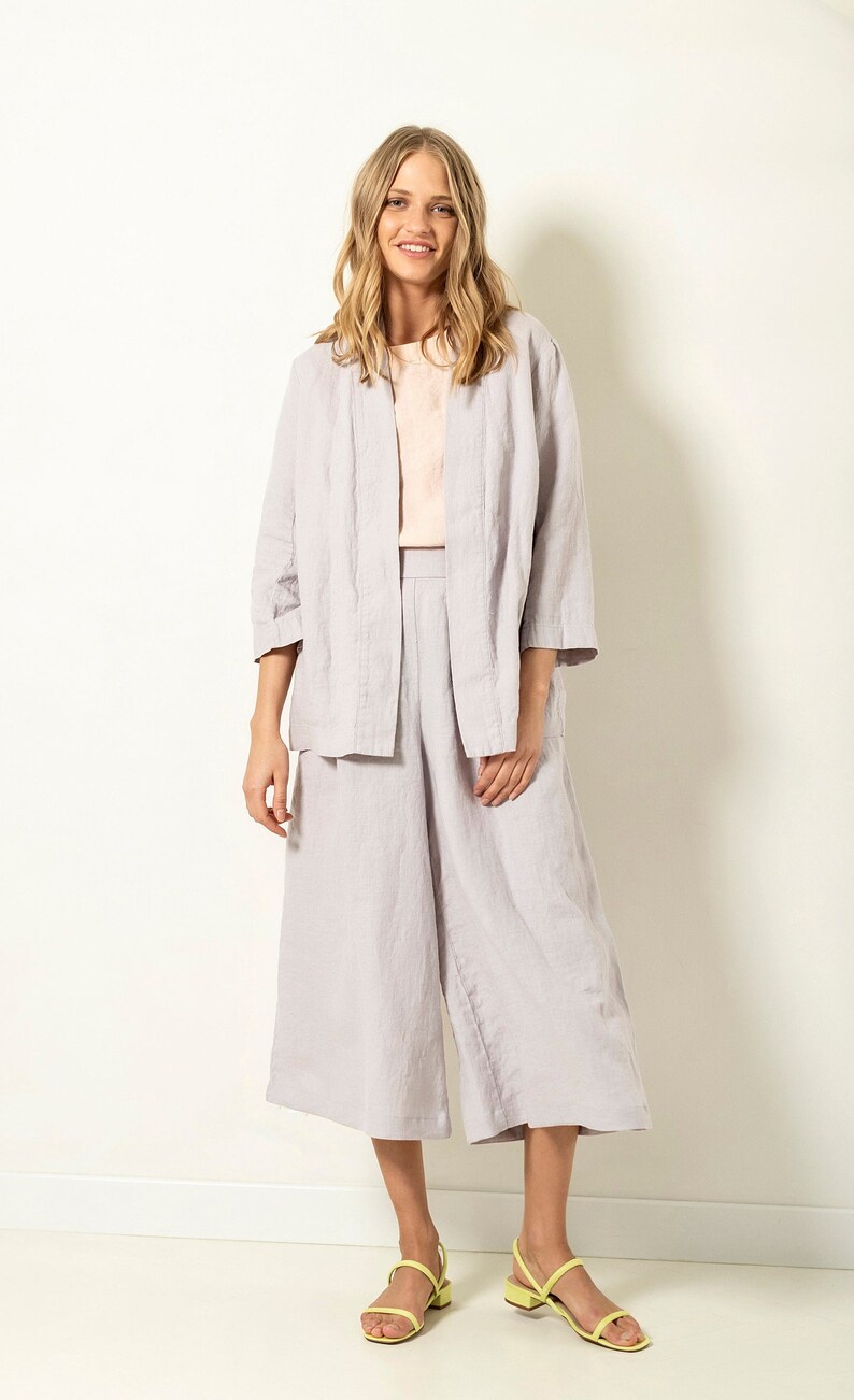 May include: A woman wearing a light purple linen outfit, consisting of a jacket, a top, and culottes. She is also wearing yellow sandals. The outfit is made of a soft, flowing fabric, perfect for warm weather.