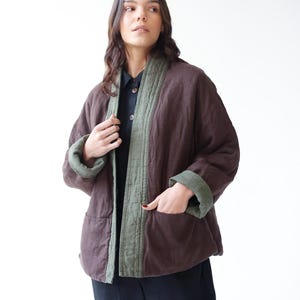 May include: A brown quilted jacket with olive green trim and cuffs. The jacket has a relaxed fit with pockets and is worn over a dark shirt and trousers. The jacket's color is a deep, earthy tone, and the trim provides a contrasting accent.