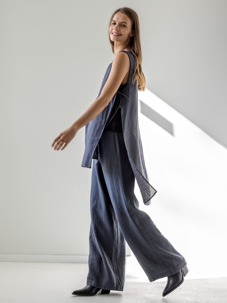Puede incluir: Una mujer lleva una blusa de lino gris oscuro y pantalones anchos a juego. La blusa tiene un corte holgado y fluido y un detalle de nudo en el lateral. Los pantalones tienen un corte holgado y una pierna ancha.