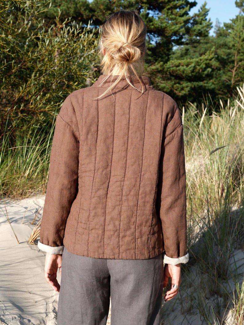 May include: A brown quilted jacket with vertical stitching, long sleeves, and a high collar. The jacket features light-colored cuffs visible at the wrists and is worn over gray trousers. The garment has a structured fit with a straight hemline.
