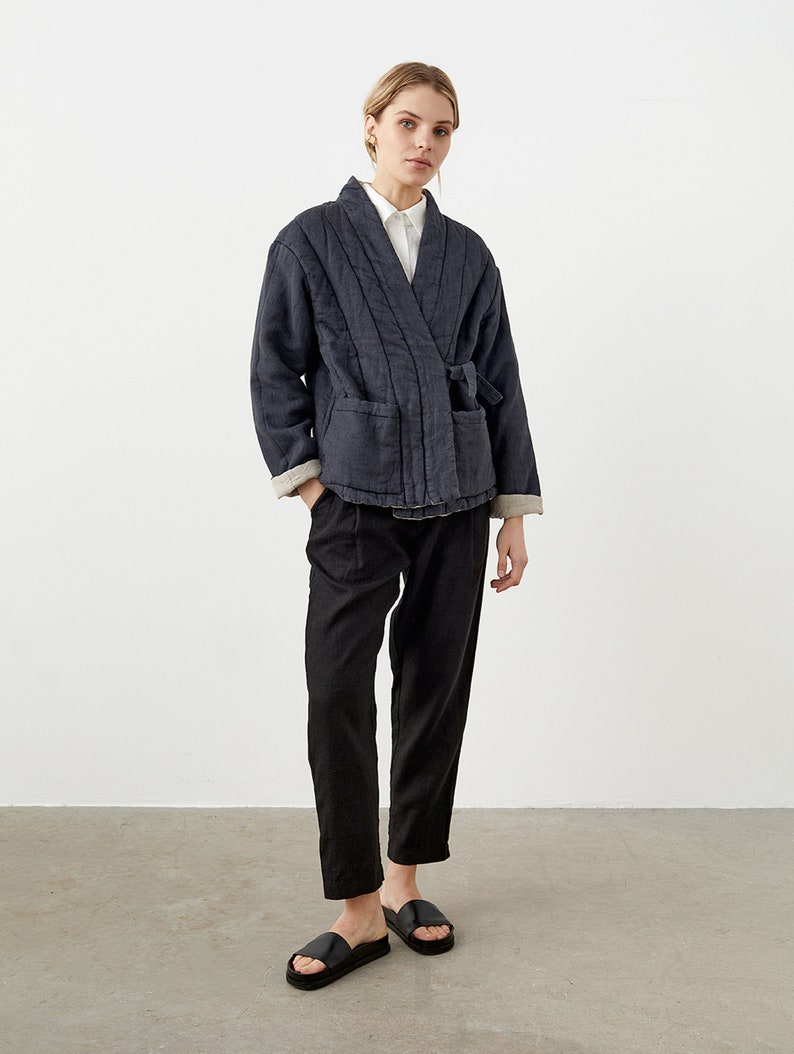 May include: A dark blue linen jacket with a wrap closure and a tie belt. The jacket has a relaxed fit and is quilted throughout. The model is wearing black pants and black sandals.
