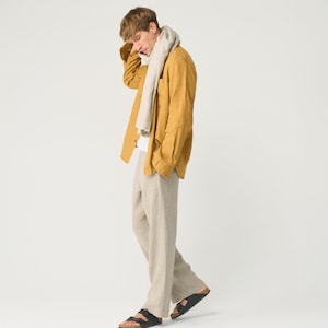 May include: A man wearing a mustard yellow linen shirt with a beige scarf, beige linen pants, and black sandals.