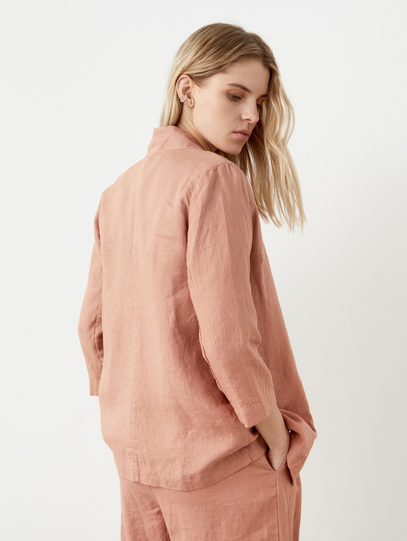 May include: A dusty rose linen blazer with three-quarter sleeves and matching pants. The blazer has a collared neckline and a relaxed fit. The outfit is made of a lightweight, textured fabric, perfect for a casual or semi-formal look.
