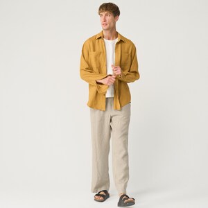 May include: A man wearing a mustard yellow linen shirt with a white t-shirt underneath and beige linen pants. He is wearing black sandals.