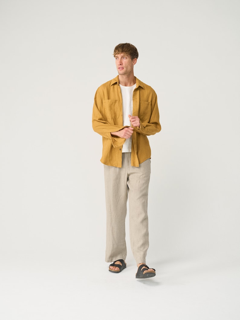 May include: A man wearing a mustard yellow linen shirt with a white t-shirt underneath and beige linen pants. He is wearing black sandals.
