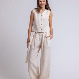 May include: A beige linen two-piece outfit. The sleeveless top has a V-neck and wooden buttons. The high-waisted, wide-leg pants have a matching fabric belt. The outfit is made of a lightweight, textured fabric, perfect for warm weather.