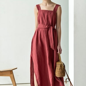 May include: A long, red linen dress with a square neckline and a self-tie belt. The dress has a side slit and is worn with a straw bag and black and white patterned shoes.