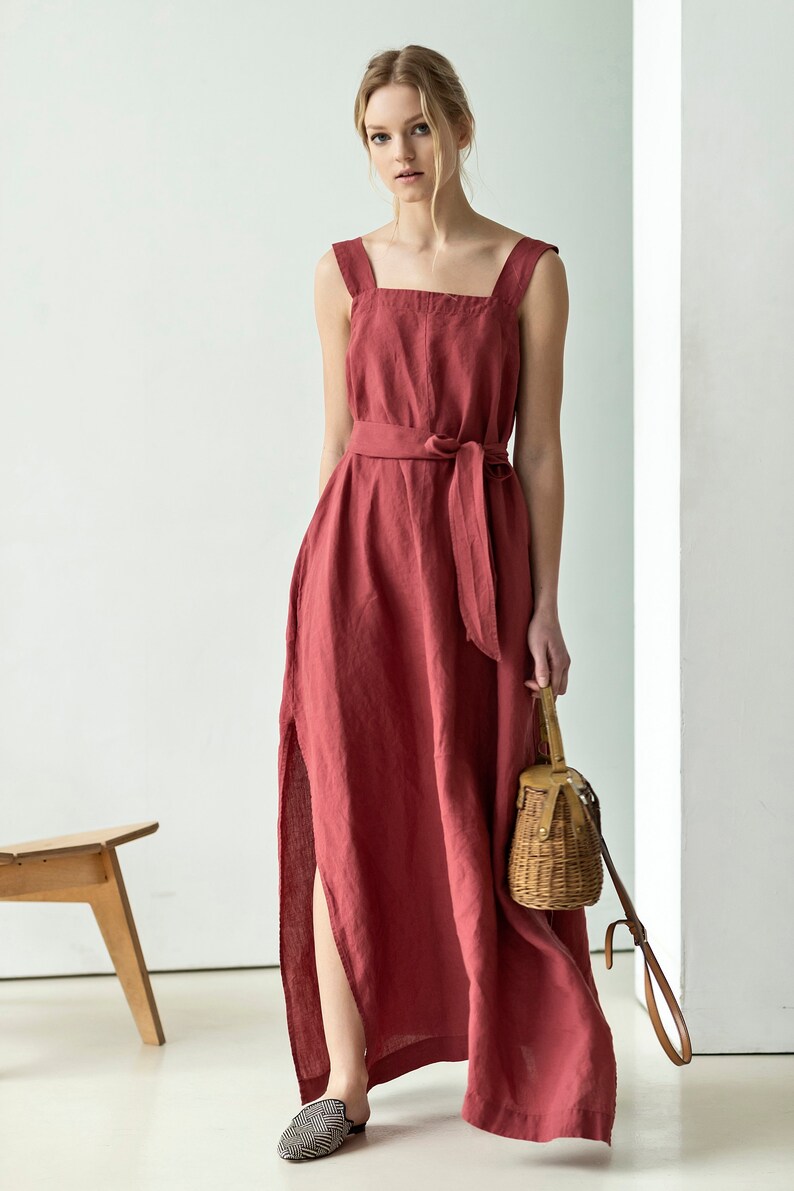 May include: A long, red linen dress with a square neckline and a self-tie belt. The dress has a side slit and is worn with a straw bag and black and white patterned shoes.