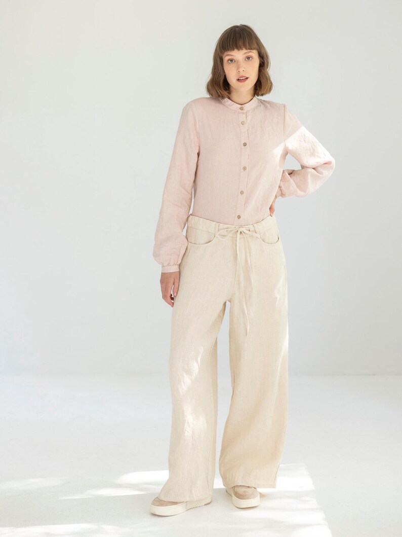 May include: A light pink long-sleeve shirt with a mandarin collar and button-down front is paired with cream-colored wide-leg pants. The pants have a drawstring waist and pockets. The outfit is completed with beige sneakers.