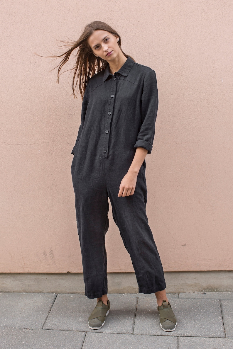 May include: A woman wearing a black linen jumpsuit with a button-up front and long sleeves. She is standing in front of a light pink wall and wearing green sneakers.