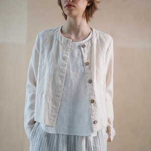 Könnte beinhalten: Hellbeige Leinenjacke mit rundem Kragen und Holzknöpfen. Die Jacke wird über einem weißen Oberteil getragen und mit einer gestreiften Hose kombiniert. Das Outfit ist schlicht und elegant, passend für Freizeitkleidung.