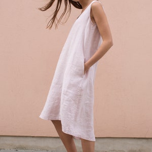 Puede incluir: Un vestido sin mangas de color rosa claro hasta la rodilla con cuello en V y bolsillos laterales, combinado con zapatillas de cordones a juego en rosa claro. La prenda presenta una silueta sencilla y holgada con un tejido texturizado.