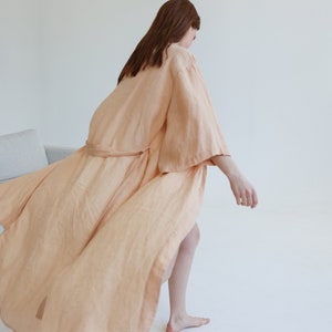 May include: A long, flowing peach-colored linen robe with a sash tie. The robe is worn by a person standing on a white background.