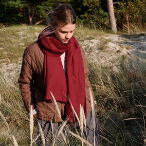 May include: A woman wearing a burgundy scarf and a brown jacket stands in a grassy field. The scarf is draped around the neck and chest. The jacket is buttoned up. The background features trees and sand.