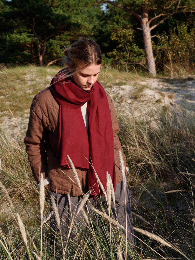 May include: A woman wearing a burgundy scarf and a brown jacket stands in a grassy field. The scarf is draped around the neck and chest. The jacket is buttoned up. The background features trees and sand.