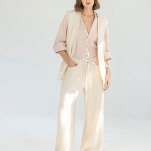 May include: A woman wearing a light pink button-down shirt, a cream-colored vest, and cream-colored wide-leg pants. The pants have a drawstring waist. The outfit is paired with beige sneakers. The background is a plain white.