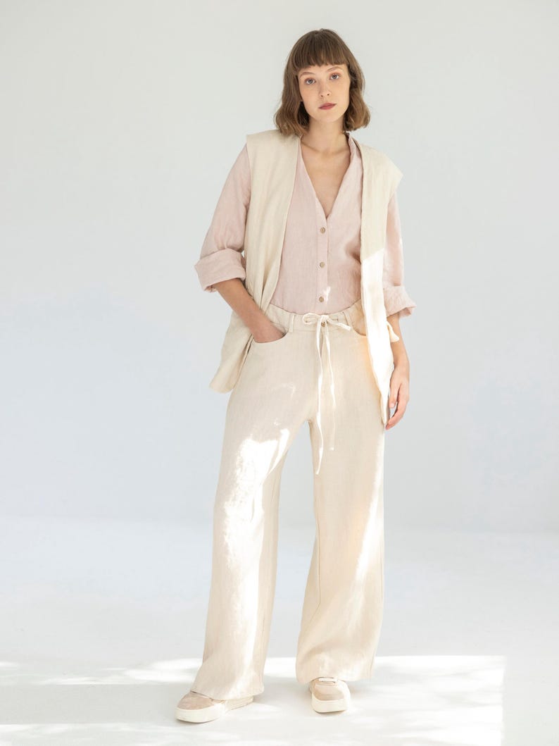 May include: A woman wearing a light pink button-down shirt, a cream-colored vest, and cream-colored wide-leg pants. The pants have a drawstring waist. The outfit is paired with beige sneakers. The background is a plain white.
