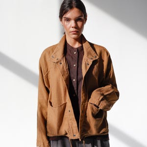 Puede incluir: Una chaqueta color canela con cuello, botones y dos bolsillos delanteros. La chaqueta se superpone a una camisa abotonada marrón oscuro y se combina con pantalones grises. El conjunto se encuentra sobre un fondo blanco con sombras.