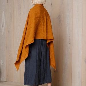 May include: An ochre-colored linen cape draped over a dark blue culotte. The outfit is paired with brown leather ankle boots with a block heel. The background is a light wood panel wall.