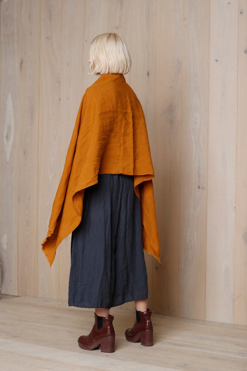 May include: An ochre-colored linen cape draped over a dark blue culotte. The outfit is paired with brown leather ankle boots with a block heel. The background is a light wood panel wall.
